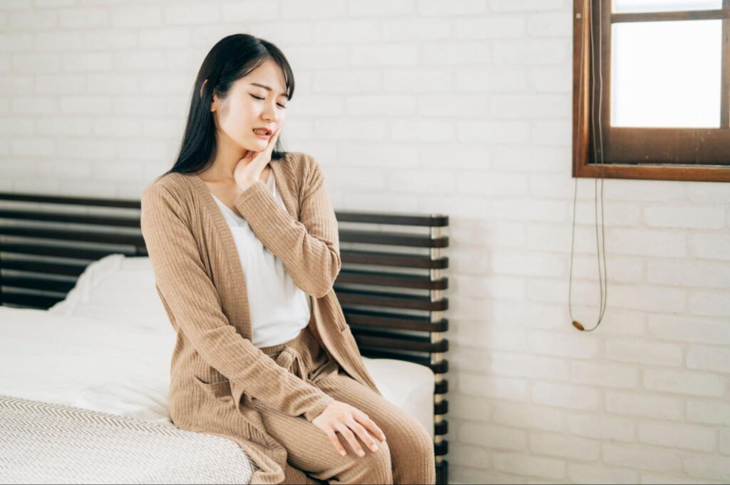 ベッドに座り親知らずに痛みを感じて頬を押さえる女性